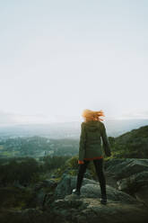 Eine einsame Abenteurerin steht auf einem Felsvorsprung auf Vancouver Island und blickt über die üppige Landschaft, während der Wind ihr Haar durcheinander wirbelt. - ADSF50838