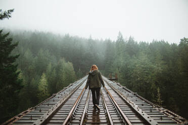 Rückenansicht einer Frau, die auf einer alten Eisenbahnstrecke durch einen nebligen Wald auf Vancouver Island spazieren geht, was ein Gefühl von Abenteuer und Geheimnis vermittelt. - ADSF50846