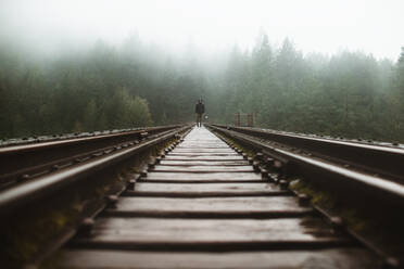 A solitary figure walks down mist-shrouded railroad tracks amidst the dense forests of Vancouver Island, evoking a sense of adventure and solitude. - ADSF50847