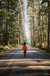 Person in orangefarbenem Pullover und Hut bei einem ruhigen Spaziergang auf einem von hohen Bäumen gesäumten Waldweg in Avatar Grove, Vancouver Island. - ADSF50856