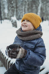 Lächelnder Junge mit Teetasse im Winterpark sitzend - ANAF02615
