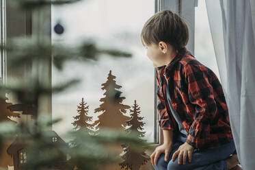 Boy near Christmas decoration looking through window at home - ANAF02619