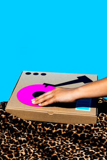 Crop hand of anonymous woman playing antique turntable made with cardboard box on yellow table over animal print textile print on blue background - ADSF50866