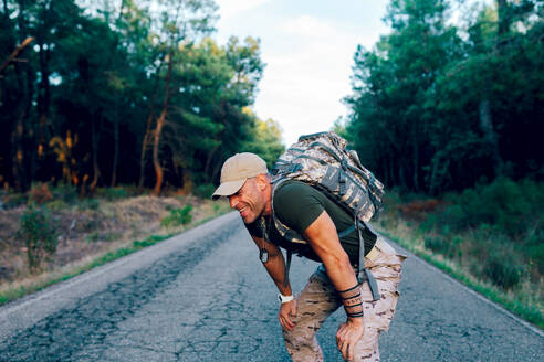 Müder reifer Marinesoldat in Uniform und mit Rucksack, der sich beim Training im Wald auf die Knie stützt und lächelt - ADSF50873