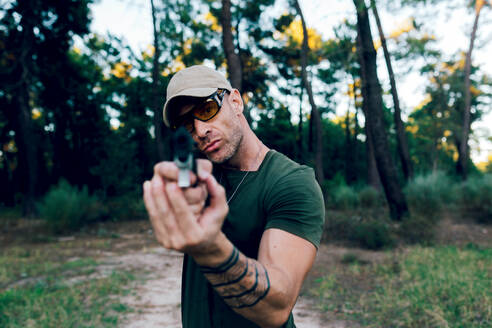 Kommandeur der Spezialeinheiten mit Mütze und Brille im mittleren Alter schaut in die Kamera, während er mit einem Gewehr im Wald gegen den Sonnenuntergang zielt - ADSF50875
