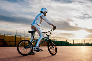 Ganzkörper-Seitenansicht eines anonymen Mannes mit Helm und in silbernem Kostüm, der wegschaut, während er auf leerem Boden gegen einen bewölkten, sonnenuntergangsblauen Himmel Fahrrad fährt - ADSF50881