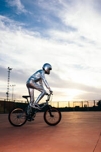 Ganzkörper-Seitenansicht eines anonymen Mannes mit Helm und in silbernem Kostüm, der wegschaut, während er auf leerem Boden Fahrrad fährt und Kunststücke gegen den wolkenverhangenen blauen Sonnenuntergangshimmel macht - ADSF50883
