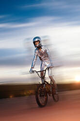 Tiefwinklige Ganzkörper-Seitenansicht eines Mannes mit Helm und in silbernem Kostüm, der wegschaut, während er auf leerem Boden Fahrrad fährt und Kunststücke gegen den bewölkten blauen Himmel bei Sonnenuntergang macht - ADSF50884