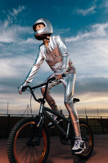 Tiefwinklige Ganzkörper-Seitenansicht eines Mannes mit Helm und in silbernem Kostüm, der wegschaut, während er auf leerem Boden Fahrrad fährt und Kunststücke gegen den bewölkten blauen Himmel bei Sonnenuntergang macht - ADSF50885
