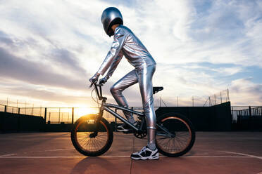 Ganzkörper-Seitenansicht eines anonymen Mannes mit Helm und in silbernem Kostüm, der wegschaut, während er auf leerem Boden Fahrrad fährt und Kunststücke gegen den wolkenverhangenen blauen Sonnenuntergangshimmel macht - ADSF50887