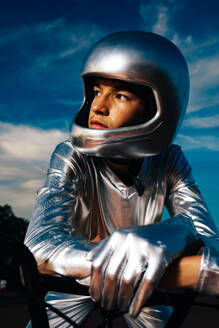 Confident young male biker in helmet and silver costume with gloves looking away while sitting on seat of bicycle and leaning on handlebar against cloudy sunset blue sky - ADSF50890