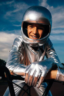 Confident young male biker in helmet and silver costume with gloves looking at camera while sitting on seat of bicycle and leaning on handlebar against cloudy sunset blue sky - ADSF50891