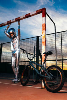 Low angle of anonymous young male in silver costume and protective helmet looking down while standing on sports ground with bicycle and holding goal post with hands against cloudy sunset blue sky - ADSF50892