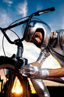 Side view of confident anonymous young male rider in helmet and silver costume with gloves looking down while kneeling near bicycle and repairing wheel brake against sunset orange sun and cloudy blue sky - ADSF50893