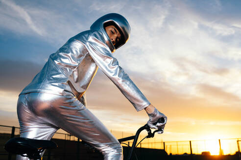 Full body side view of male in helmet and in silver costume looking at camera while riding bicycle on empty ground and doing tricks against cloudy sunset blue sky - ADSF50895