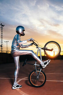 Ganzkörper-Seitenansicht eines anonymen Stuntman in silbernem Kostüm und Helm, der wegschaut, während er stehend den Lenker mit dem Bein auf dem Pedal hält und das Fahrrad auf dem Hinterrad auf einem Sportplatz vor einem orangefarbenen Sonnenuntergang steht - ADSF50896