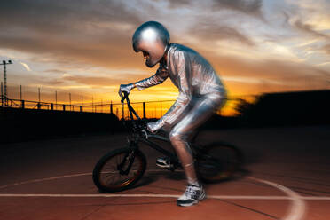 Ganzkörper-Seitenansicht eines anonymen Mannes mit Helm und in silbernem Kostüm, der wegschaut, während er auf leerem Boden Fahrrad fährt und Kunststücke gegen den wolkenverhangenen blauen Sonnenuntergangshimmel macht - ADSF50897