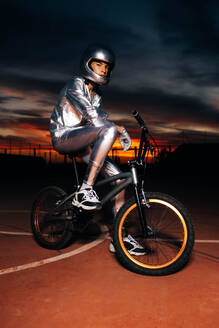 Side view of young anonymous stuntman in silver costume and helmet looking at camera while sitting on bicycle seat with sneakers foot on sports ground against dark cloudy orange sky in light - ADSF50899