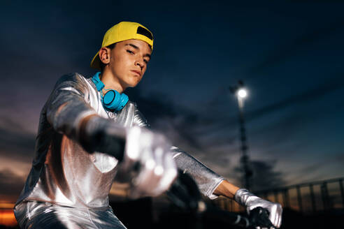 Low angle of young male in silver costume with gloves and wearing yellow cap looking at camera while riding bicycle with headphones on neck and holding handlebar against dark cloudy evening sky in light - ADSF50900