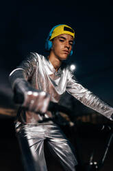 Niedriger Winkel eines jungen Mannes im silbernen Kostüm mit Handschuhen und gelber Mütze, der in die Kamera schaut, während er mit Kopfhörern am Ohr Fahrrad fährt und den Lenker gegen einen dunklen, bewölkten Abendhimmel hält - ADSF50901