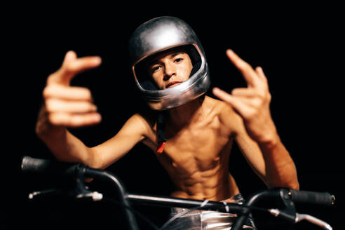 Anonymous shirtless young stuntman in helmet looking at camera while sitting on bicycle seat with fingers displaying rock and role gesture with elbows on handlebar in illuminated light against dark background - ADSF50903