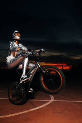 Seitenansicht eines anonymen männlichen Motorradfahrers mit Schutzhelm, der wegschaut, während er auf dem Fahrradsattel sitzt und das Vorderrad über den Sportplatz zieht, vor einem orangefarbenen, dunklen Himmel bei Sonnenuntergang - ADSF50904