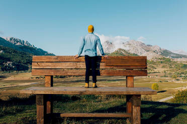 Ein Mann steht auf einer großen Holzbank mit Blick auf eine atemberaubende Bergkette unter einem strahlend blauen Himmel und verkörpert den Geist von Abenteuer und Gelassenheit. - ADSF50907