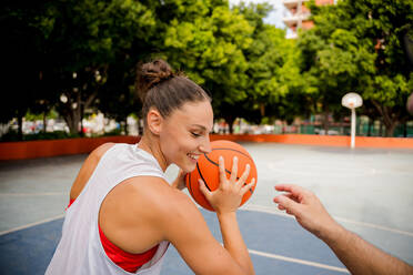 Lächelnde fitte junge Frau in Sportkleidung, die einen Basketball hält, während sie mit einem männlichen Freund auf einem Spielplatz mit Bäumen im Hintergrund spielt - ADSF50914