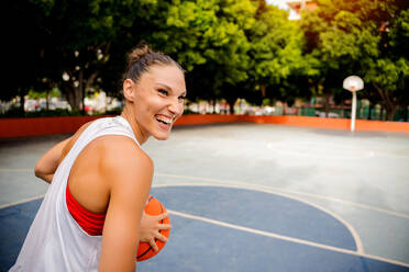 Fröhliche junge Frau in Sportkleidung, die einen Ball hält und wegschaut, während sie auf einem Spielplatz mit grünen Bäumen im Hintergrund Basketball spielt - ADSF50915