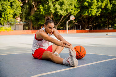 Ganzer Körper einer entschlossenen jungen Basketballspielerin, die sitzt und die Füße berührt, während sie auf dem Spielplatz Aufwärmübungen macht - ADSF50916