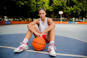 Ganzkörperaufnahme einer sportlichen jungen Frau in Sportkleidung, die mit einem Ball sitzt und in die Kamera schaut, auf einem Basketballplatz mit unscharfen Bäumen im Hintergrund - ADSF50917