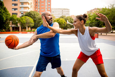 Entschlossener Mann und Frau in Sportkleidung spielen Basketball auf einem Spielplatz in einer Stadt mit Gebäuden und Bäumen im Hintergrund - ADSF50918