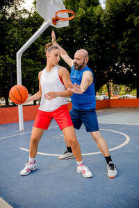Entschlossener Mann und Frau in Sportkleidung spielen Basketball auf einem Spielplatz in einer Stadt mit Gebäuden und Bäumen im Hintergrund - ADSF50919