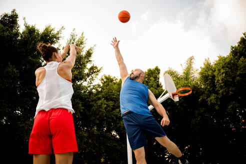 Von unten Sportlerin in Activewear wirft Basketball in Richtung Korb und Mann versucht, auf dem Platz gegen den Himmel zu verteidigen - ADSF50920