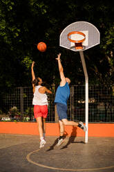 Full length back view of sportswoman in activewear shooting basketball towards basket and man trying to defend on court against sky - ADSF50923