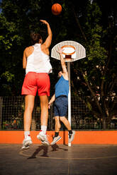 Full length back view of sportswoman in activewear shooting basketball towards basket and man trying to defend on court against sky - ADSF50924
