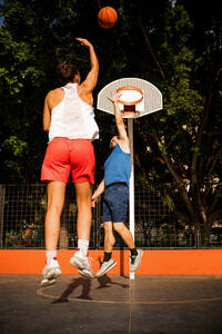 Rückenansicht einer Sportlerin in Sportkleidung, die einen Basketball in Richtung Korb schießt, und eines Mannes, der versucht, sich auf dem Platz gegen den Himmel zu verteidigen - ADSF50924