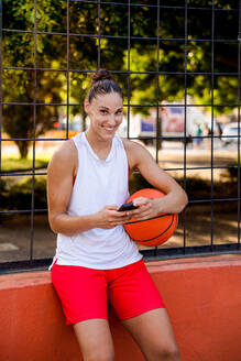 Lächelnde junge Basketballspielerin, die einen Ball hält und ein Smartphone benutzt, während sie in die Kamera auf einem Spielplatz an einem sonnigen Tag schaut - ADSF50925