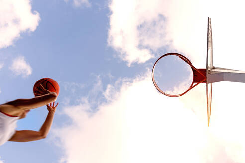 Ausschnitt von unten einer nicht erkennbaren weiblichen Basketballspielerin in Sportkleidung, die springt und den Ball in einen Korb vor einem bewölkten Himmel wirft - ADSF50926