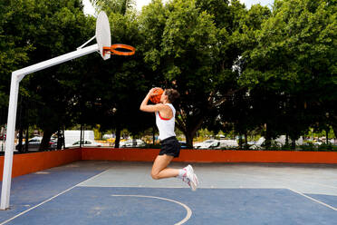 Full length side view of determined young female in sportswear jumping and throwing ball to make basket while playing on court - ADSF50928