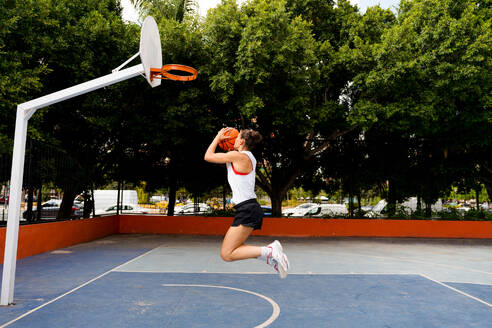 Seitenansicht in voller Länge einer entschlossenen jungen Frau in Sportkleidung, die springt und den Ball wirft, um einen Korb zu machen, während sie auf dem Platz spielt - ADSF50928