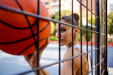 Confident young female athlete in sportswear holding basketball looking at camera seen through fence of playground - ADSF50931