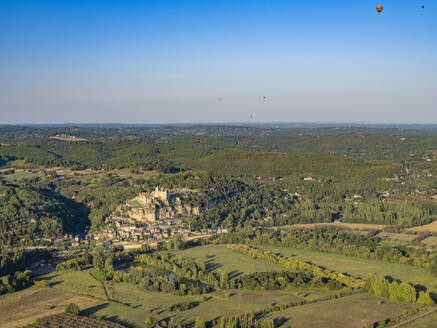 Luftaufnahme des Dorfes La Roque-Gageac und der Burg auf dem Hügel entlang der Dordogne mit Heißluftballons am Himmel, Nouvelle-Aquitaine, Südwestfrankreich - AAEF25037