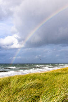 Deutschland, Mecklenburg-Vorpommern, Grasbewachsener Strand mit Regenbogen vor wolkenverhangenem Himmel über dem Meer im Hintergrund - EGBF00996