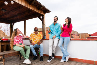 Multiethnische Gruppe von glücklichen jungen Freunden mit Abendessen Grillparty auf dem Dach zu Hause - Multirassische fröhliche junge erwachsene Menschen, die Spaß haben und sich auf einer Terrasse Balkon mit Blick auf die Stadt, Essen und Trinken. - DMDF08541