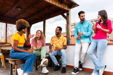 Multiethnische Gruppe von glücklichen jungen Freunden mit Abendessen Grillparty auf dem Dach zu Hause - Multirassische fröhliche junge erwachsene Menschen, die Spaß haben und sich auf einer Terrasse Balkon mit Blick auf die Stadt, Essen und Trinken. - DMDF08542
