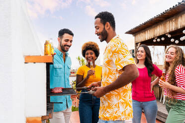 Multiethnische Gruppe von glücklichen jungen Freunden mit Abendessen Grillparty auf dem Dach zu Hause - Multirassische fröhliche junge erwachsene Menschen, die Spaß haben und sich auf einer Terrasse Balkon mit Blick auf die Stadt, Essen und Trinken. - DMDF08556