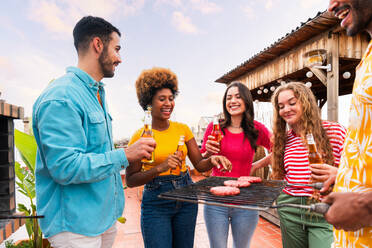 Multiethnische Gruppe von glücklichen jungen Freunden mit Abendessen Grillparty auf dem Dach zu Hause - Multirassische fröhliche junge erwachsene Menschen, die Spaß haben und sich auf einer Terrasse Balkon mit Blick auf die Stadt, Essen und Trinken. - DMDF08557