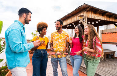 Multiethnische Gruppe von glücklichen jungen Freunden mit Abendessen Grillparty auf dem Dach zu Hause - Multirassische fröhliche junge erwachsene Menschen, die Spaß haben und sich auf einer Terrasse Balkon mit Blick auf die Stadt, Essen und Trinken. - DMDF08559