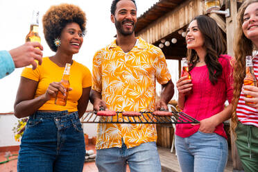 Multiethnische Gruppe von glücklichen jungen Freunden mit Abendessen Grillparty auf dem Dach zu Hause - Multirassische fröhliche junge erwachsene Menschen, die Spaß haben und sich auf einer Terrasse Balkon mit Blick auf die Stadt, Essen und Trinken. - DMDF08560